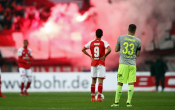 Kommerzialisierung und Ultras: Was wird aus dem Fußball? MAINZ, GERMANY - NOVEMBER 18: Yoshinori Muto #9 of Mainz and Matthias Lehmann of Koeln react as fans of Koeln burn flares during the Bundesliga match between 1. FSV Mainz 05 and 1. FC Koeln at Opel Arena on November 18, 2017 in Mainz, Germany. (Photo by Alex Grimm/Bongarts/Getty Images)